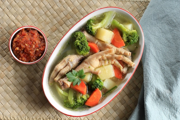 Photo clear chicken feet (claw) soup with potato, broccoli, and carrots. served on marble table in brown bowl with sambal