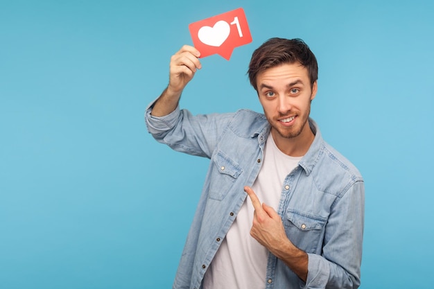 Click Like. Portrait of cheerful handsome blogger man in denim shirt pointing at social media heart Like button, emoji counter, follower notification. indoor studio shot isolated on blue background