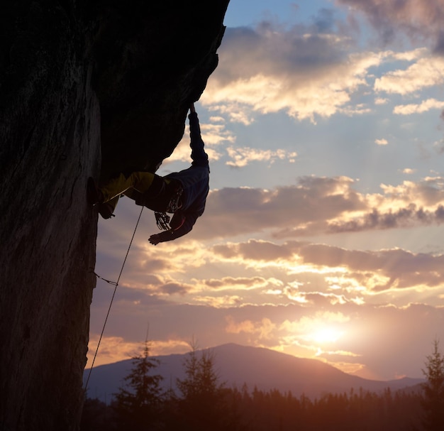 Climber holding with one hand on hanging rock at sunrise in mountains Extreme rock climbing Cloudy sky with copy space