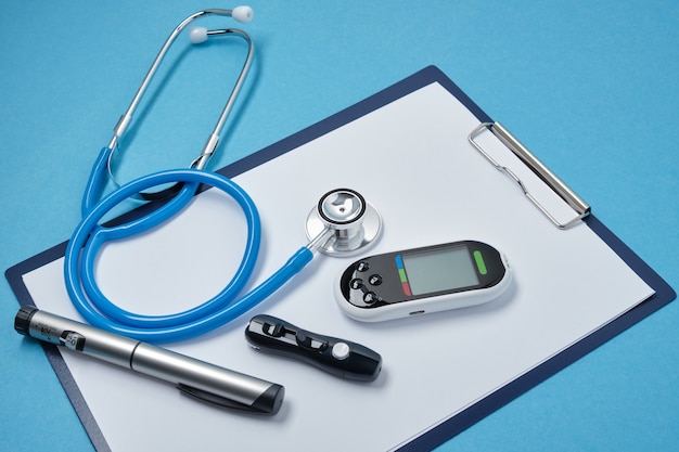 Clipboard with white blank sheets of paper, stethoscope, glucose meter, lancet and syringe pen with insulin on blue background, daibet day concept, diabetes diagnostics