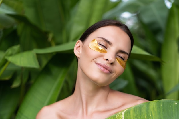 Close beauty portrait of a topless woman with perfect skin and natural makeup shiny patches under the eyes to moisturize the skin posing in tropical green leaves outdoor