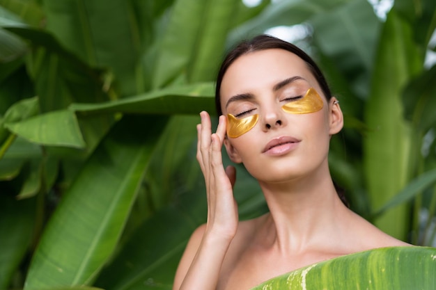 Close beauty portrait of a topless woman with perfect skin and natural makeup shiny patches under the eyes to moisturize the skin posing in tropical green leaves outdoor