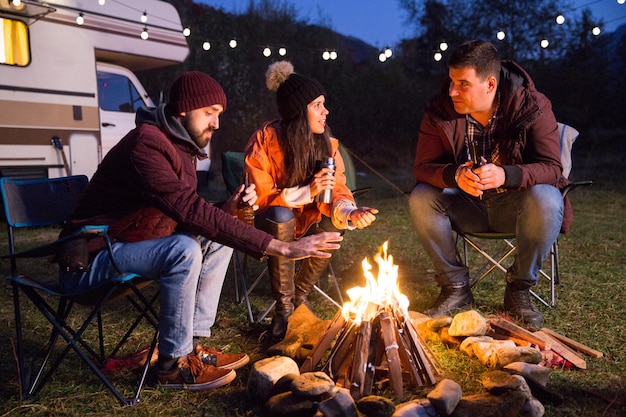 Close friends ejoying the heat from their camp fire in a cold night of autumn in the mountains. Retro camper van with light bulbs.