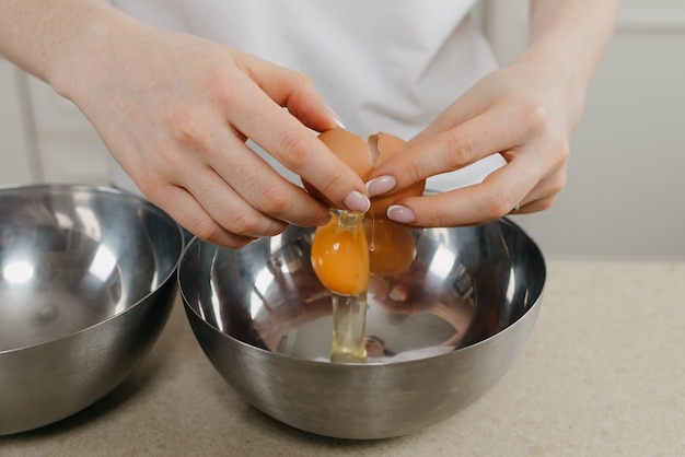 A close photo of the hands of a young woman breaking egg above the stainless steel bowl in the kitchen