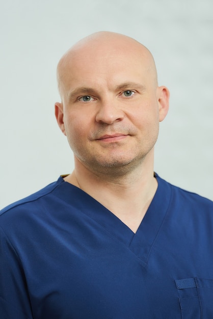 A close portrait of a caucasian bald serious adult dentist in the navy blue uniform is posing in a dentist's office