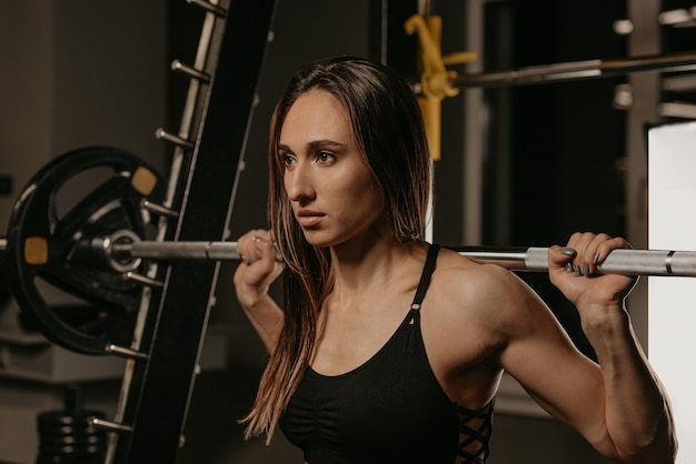 A close portrait of a sporty woman with dark hair who is lifting a barbell in the squat rack in a gym. A girl is doing a leg workout.