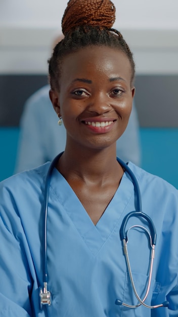 Close up of african american nurse using laptop computer on white desk at healthcare clinic. Black medical assistant typing on device while looking at camera with stethoscope and uniform