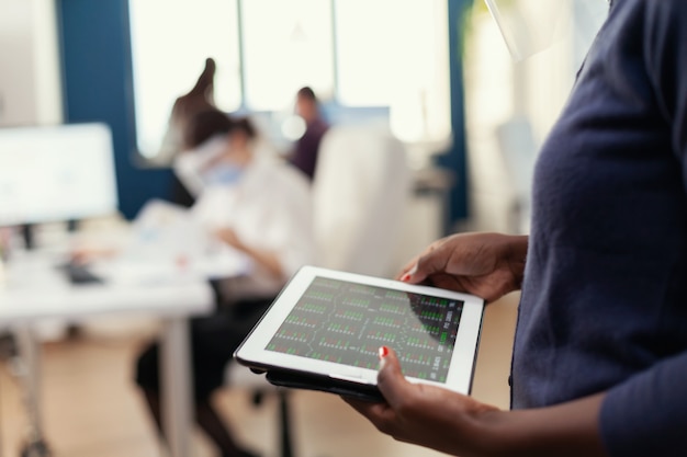 Close up of african entrepreneur in busy office using tablet pc analysing financial graph. Multiethnic coworkers respecting social distance in business company during global pandemic with coronavirus.