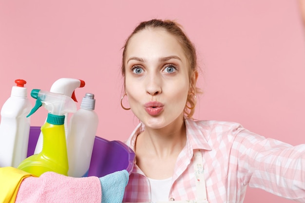 Close up of amazed woman housewife in apron hold basin with detergent bottles washing cleansers doing housework isolated on pink background. Housekeeping concept. Doing selfie shot on mobile phone.