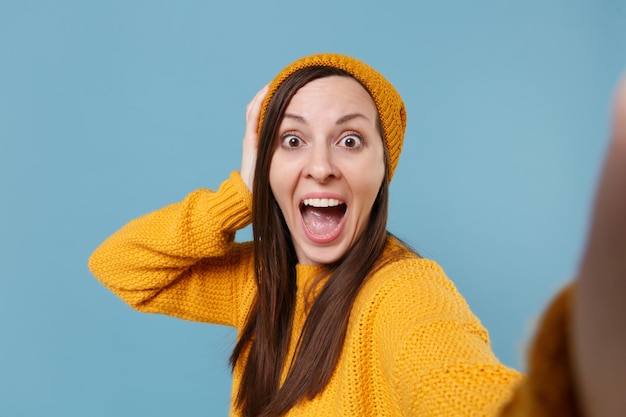 Close up of amazed young woman in sweater and hat posing isolated on blue background studio portrait. People lifestyle concept. Mock up copy space. Doing selfie shot on mobile phone put hand on head.