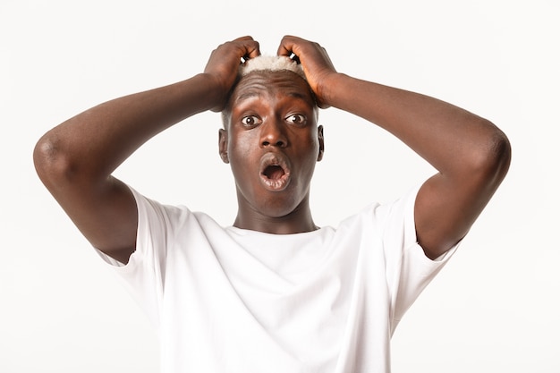 Close-up of ambushed and shocked african-american blond guy, gasping and looking startled, holding hands on head