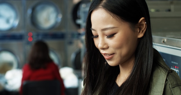 Close up of Asian pretty woman with long dark hair tapping and texting message on smarphone while standing in laundry service room. Beautiful woman typing on phone and waiting for clothes to wash.