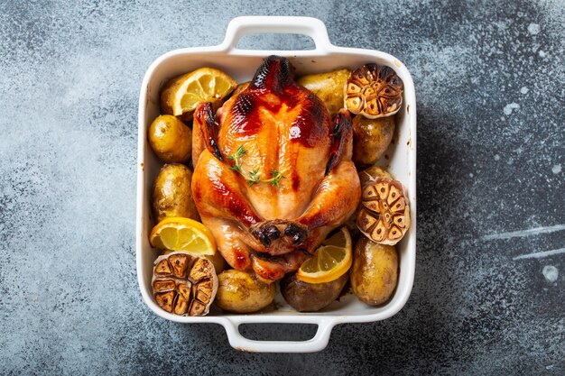 Close-up of baked or roasted whole juicy crispy organic chicken with potatoes, garlic, lemon in white casserole dish on concrete rustic background, top view