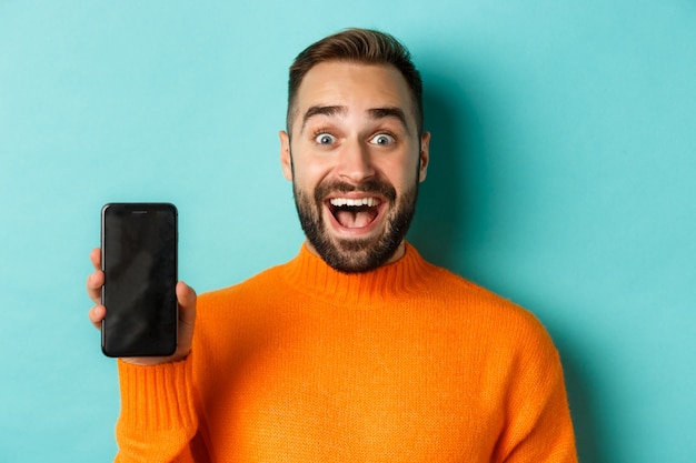 Close-up of  bearded guy in orange sweater, showing smartphone screen and smiling, showing promo online