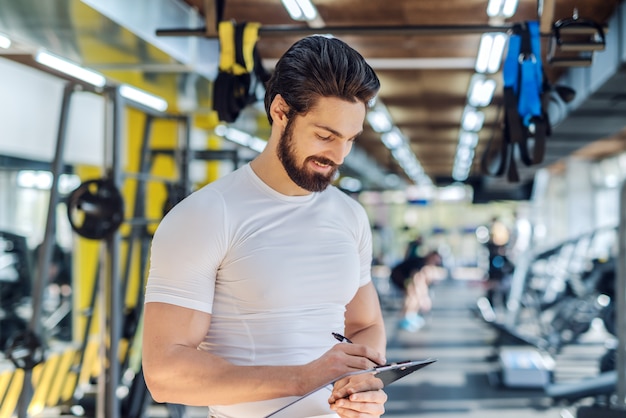 Close up of bearded personal trainer writing on clipboard results of training. Gym interior.