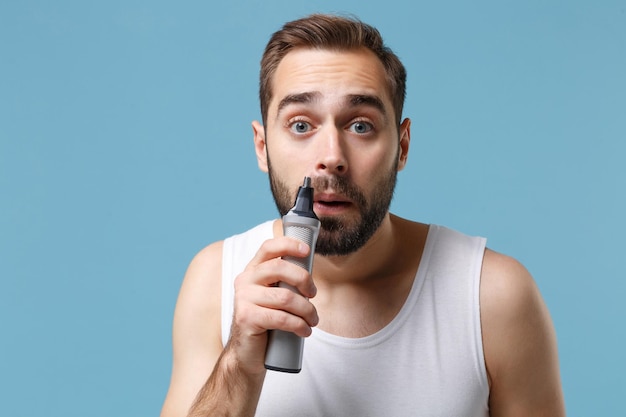 Close up bearded young man 20s years old in white shirt hold electric trimmer isolated on blue pastel background, studio portrait. Skin care healthcare cosmetic procedures concept. Mock up copy space.