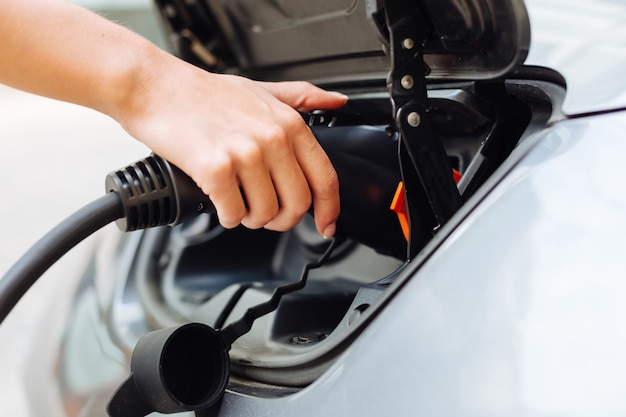 The close up of beautiful delicate female hands holding an electrical nozzle and charging an electric car