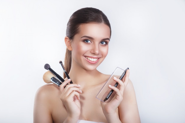 Close-up of beautiful young smiling female model with cosmetic brushes and eye shadow on white wall.