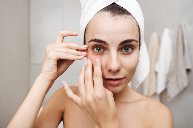 Close up of a beautiful young woman with towel on her head standing at the bathroom, applying eye patches