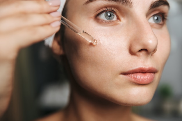 Close up of a beautiful young woman wrapped in towel applying cosmetic oil on her face