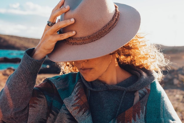 Close up of beauty portrait of woman hidden face under a traveler hat Outdoor leisure activity female with trendy clothes Concept of travel lifestyle people Pretty lady model outdoor in sunset time