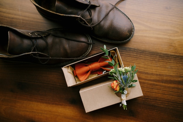 Close-up of a bow tie for a groom with a boutonniere on a cardboard box and men's shoes on a brown floor