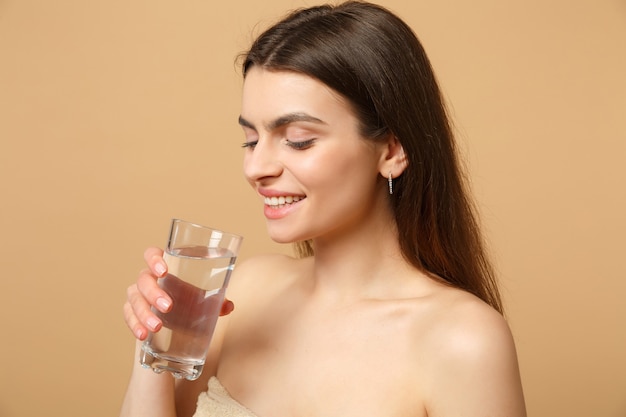 Close up brunette half naked woman with perfect skin nude make up glass water isolated on beige pastel wall