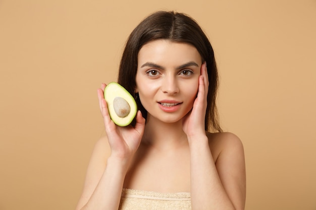 Close up brunette half naked woman with perfect skin nude make up holds avocado isolated on beige pastel wall