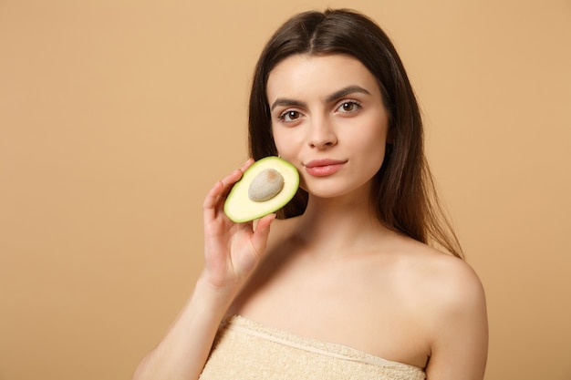 Close up brunette half naked woman with perfect skin nude make up holds avocado isolated on beige pastel wall