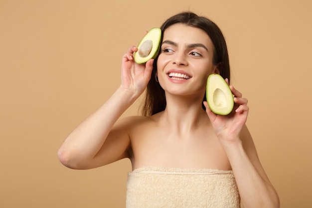 Close up brunette half naked woman with perfect skin nude make up holds avocado isolated on beige pastel wall