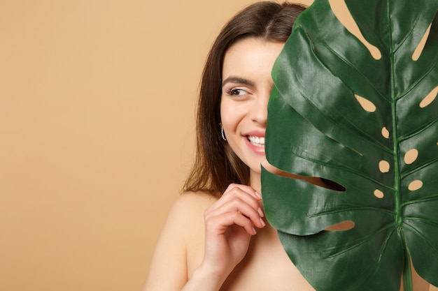 Close up brunette half naked woman with perfect skin, nude make up and palm leaf isolated on beige pastel wall