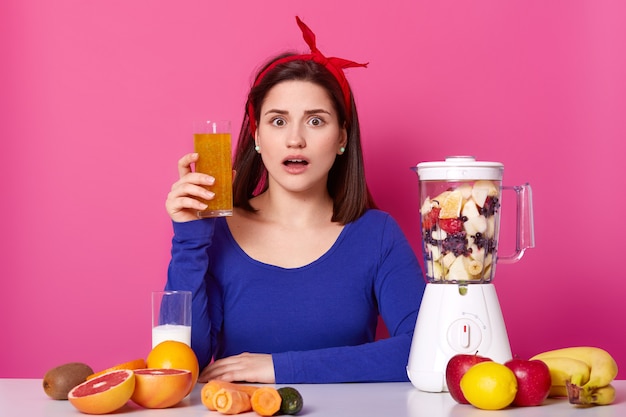 Close up of brunette woman with glass of fresh orange juice in hand. Lady likes healthy eating, making smoothie at home. Surprised female with blender against pink wall. Dieting and people concept.