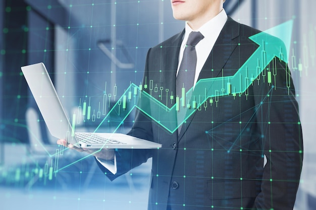 Close up of businessman hand holding laptop with growing green candlestick arrow on blurry office interior background Financial growth market and trade concept Double exposure