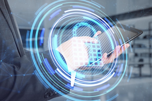 Close up of businessman hand pointing at tablet with glowing padlock hologram on blurry office interior background Secure safety and web protection concept Double exposure