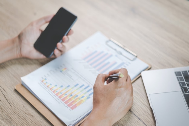 Close-up businessman holding a pen, smartphone and pointing at financial graph checking business report on wooden desk with computer laptop besided at home. Stock photo