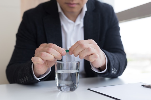 Close up of businessman in the office holding pills and glass of fresh water, taking medicine for headache, abdominal pain or taking vitamins, healthcare concept