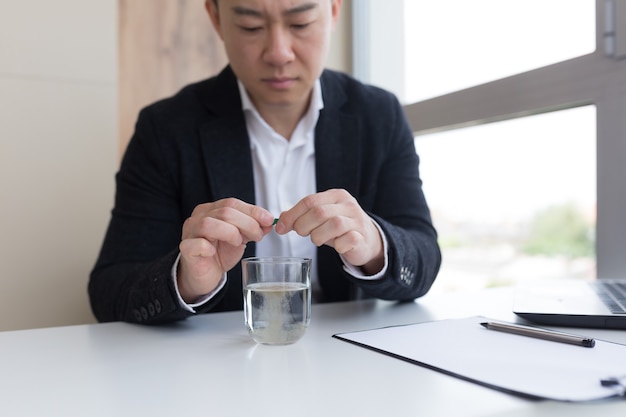 Close up of businessman in the office holding pills and glass of fresh water, taking medicine for headache, abdominal pain or taking vitamins, healthcare concept