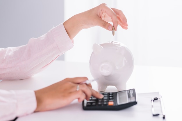 Close-up of businesswoman using calculator inserting coin in piggybank