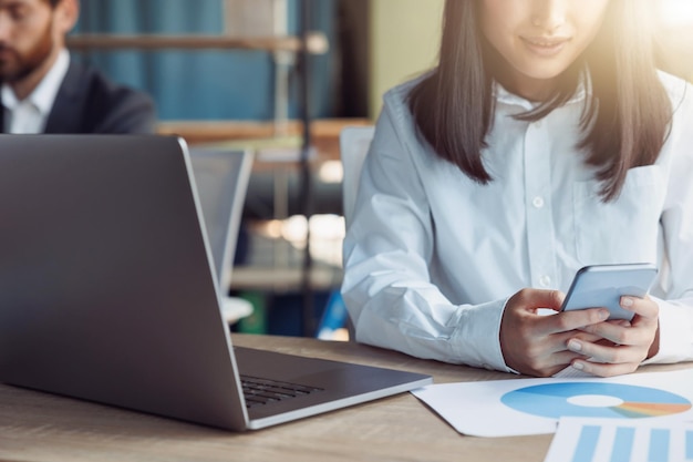 Close up of businesswoman using phone while sit on workplace in office on background of colleague