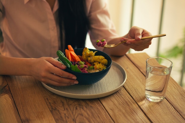 Close up of the cafe table and a glass of water with a bowl of vegetables on it. Hand of woman eating meal with a fork