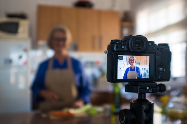 Close up of camera recording an old mature woman senior cooking and showing some recipes to her channel - youtuber job streaming in live or doing cook videos in the kitchen of her house