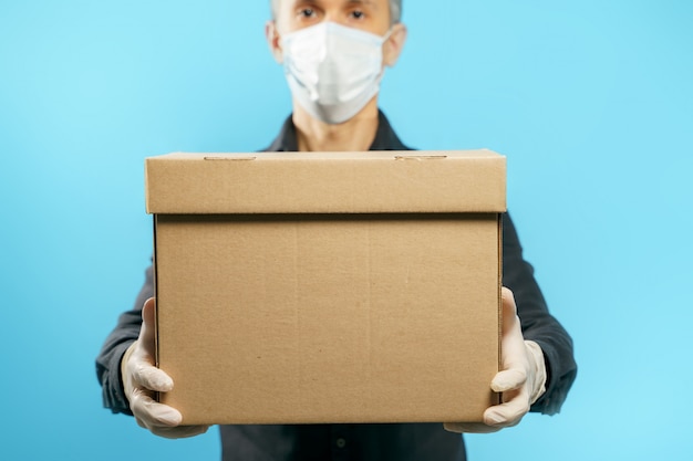 Close-up of a cardboard box in the hands of a young man in a medical protective mask and gloves on blue. Secure delivery