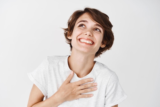 Close up of carefree young woman in tshirt holding hand on heart looking up and smiling grateful standing dreamy against white background