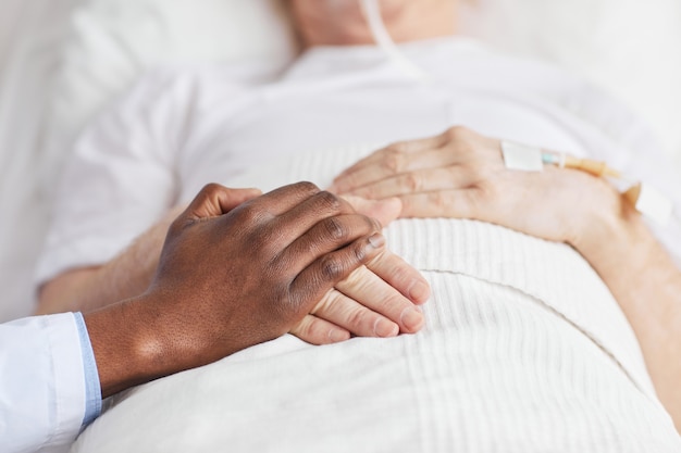 Close up of caring African-American doctor holding hands with senior patient lying in hospital bed, copy space