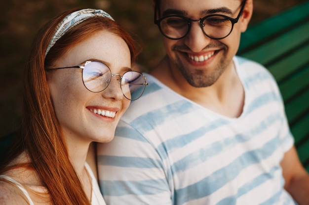 Close up of a charming young couple laughing while he is looking at her outdoor sitting in the park.