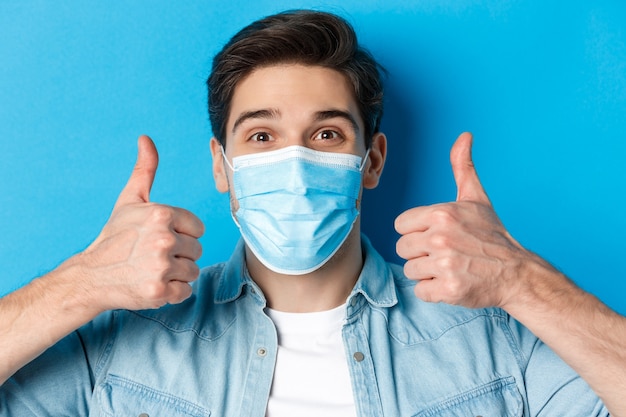 Close-up of cheerful young man in medical mask smiling, showing thumbs up in approval, like and agree, standing over blue wall