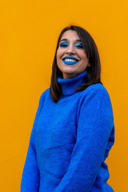 Close up of cheerful young woman with blue eye shadow makeup. Beauty portrait of female model with vivid makeup laughing on yellow background.