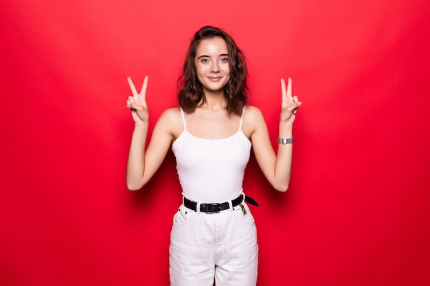 Close up of a cheery stylish girl wearing hat showing peace gesture isolated over pink wall