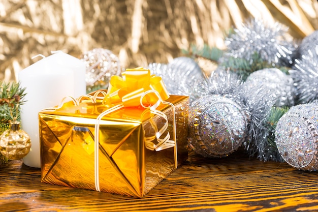 Close Up of Christmas Gift Wrapped in Gold on Rustic Wooden Table with Candle and Evergreen Branches Decorated in Silver Garland and Balls