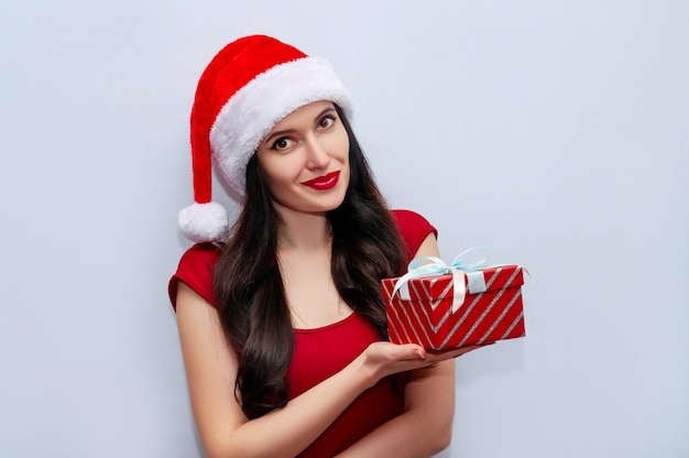 Close up Christmas Photo of Beautiful Woman with Gift Box in Red Dress and Santa Hat on Grey space.